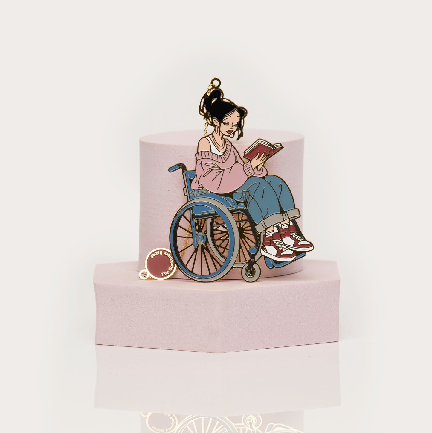 Enamel bookmark of a person in a wheelchair reading a book on a pink stand with a white background