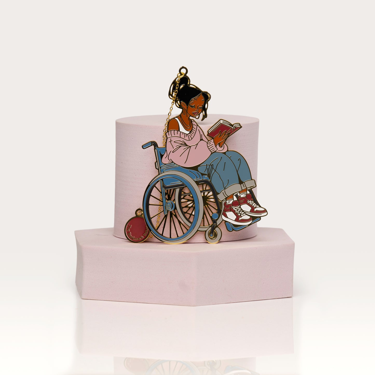 Enamel bookmark of a person in a wheelchair reading a book on a pink base with a white background