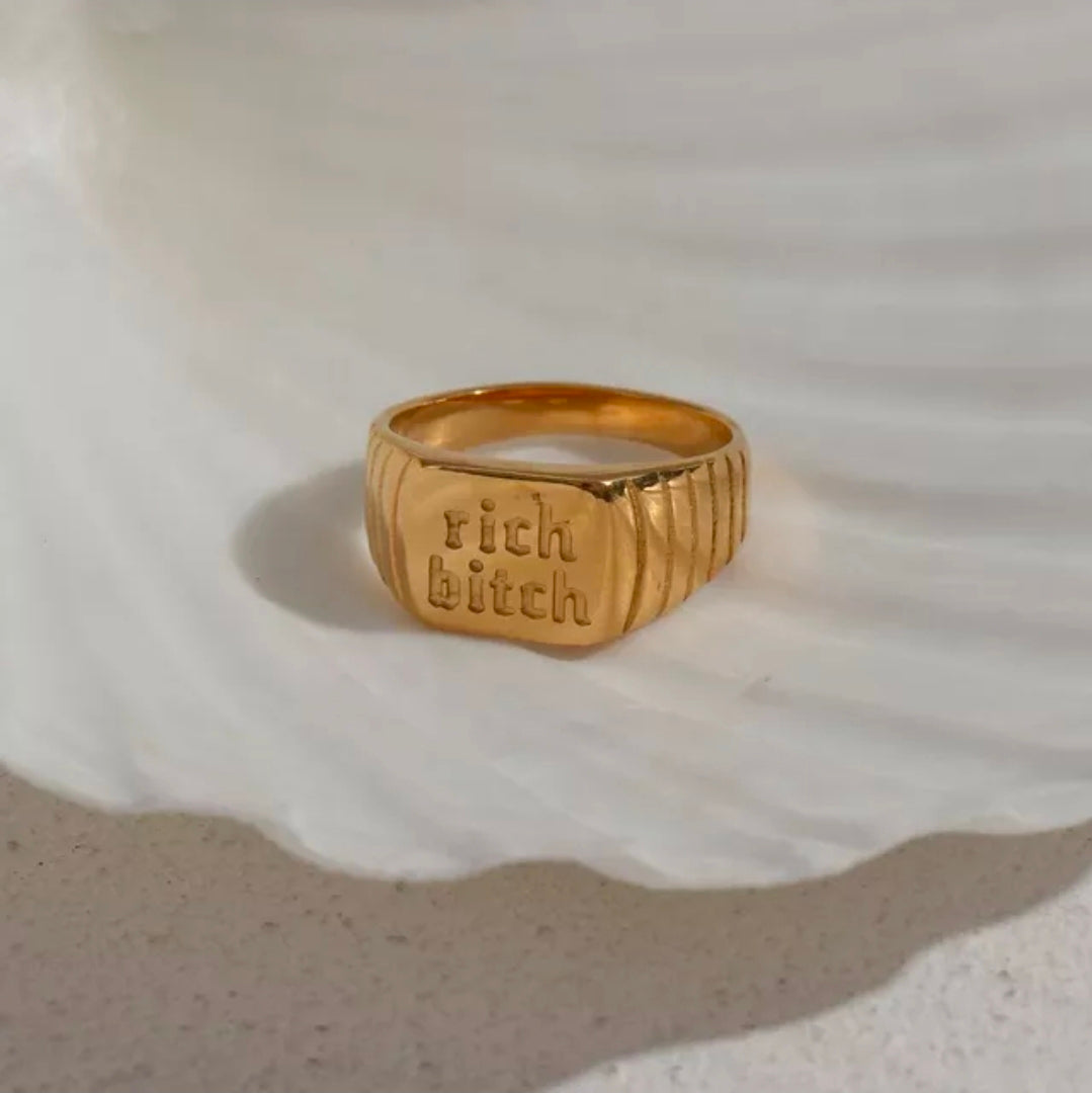 Gold statement ring engraved with “rich bitch,” styled on a neutral shell surface with a minimal, editorial aesthetic