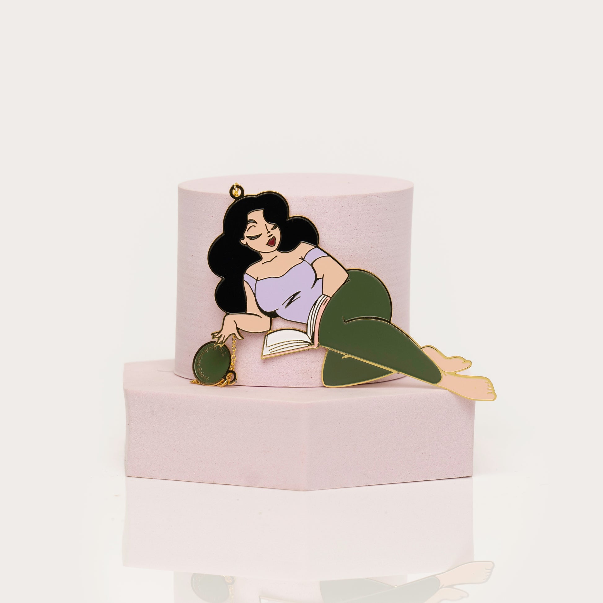 Enamel bookamark of a woman lying on a book with green pants a white background