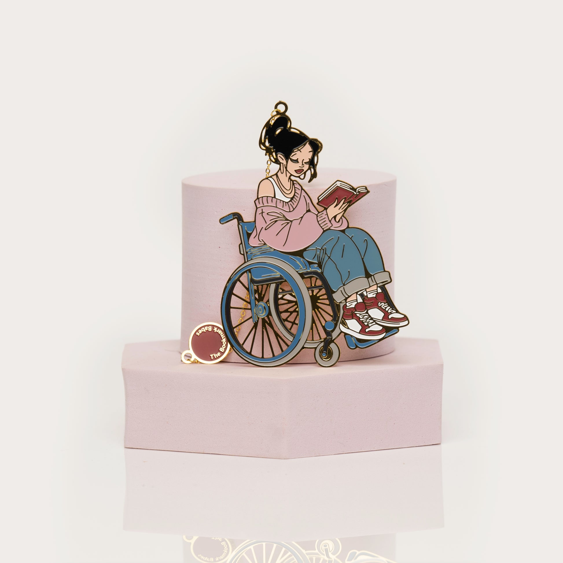 Enamel bookmark of a person in a wheelchair reading a book on a pink stand with a white background