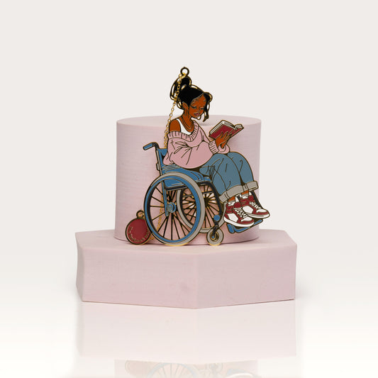 Enamel bookmark of a person in a wheelchair reading a book on a pink base with a white background