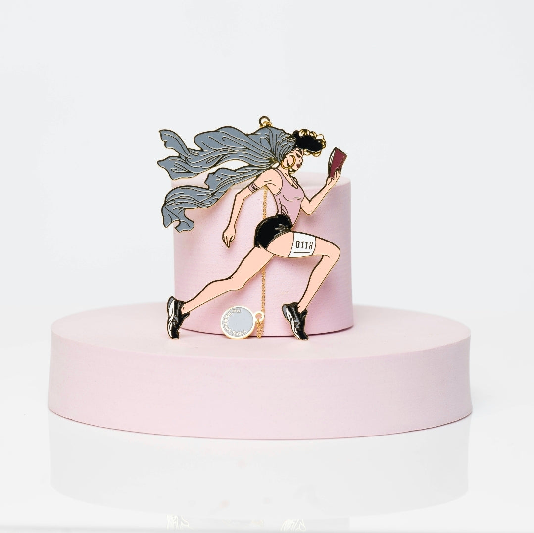 Enamel bookmark of a Black woman running with a book in hand, wearing a race bib and athletic gear, with a long gray ponytail flowing behind her