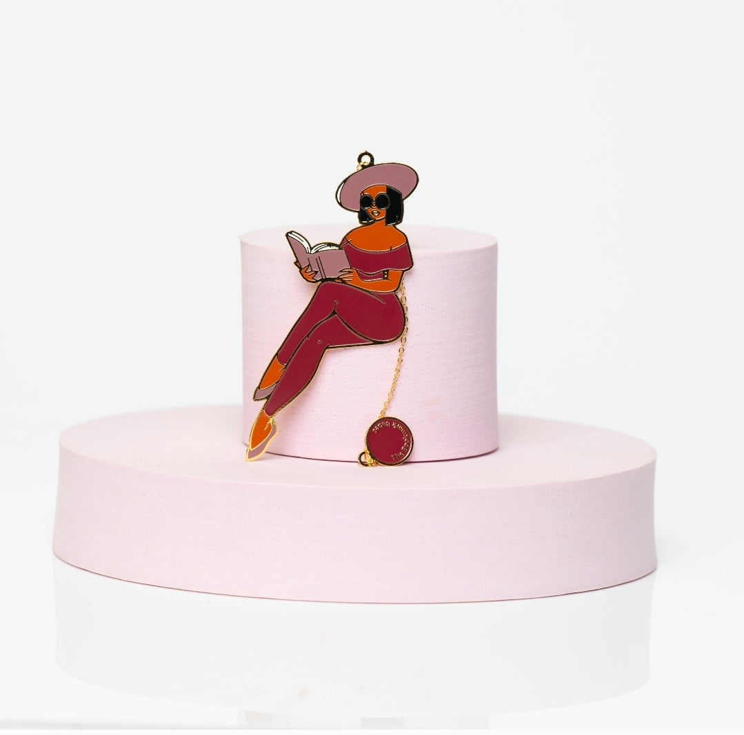 Enamel bookmark featuring Portia, a stylish Black woman in a red catsuit, reading a book with sunglasses and a wide-brim hat. Includes gold chain and charm, displayed on a pink pedestal.