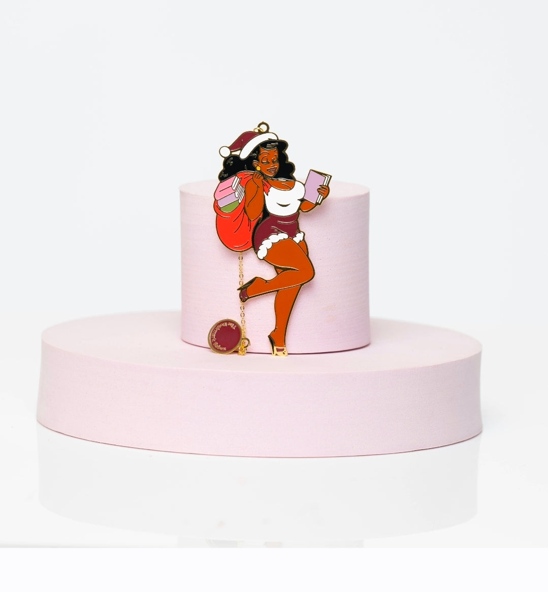 Enamel bookmark charm featuring St. Niki, a glamorous Black Santa-inspired Babe with a red gift bag, pink book, white fur-trimmed outfit, and gold heels, posing confidently on a pastel platform.