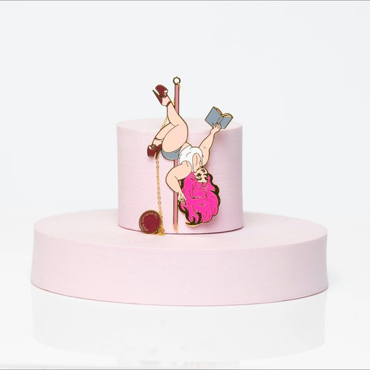 Enamel bookmark featuring Boudoir Bambi, a pink-haired pole-dancing pin-up babe reading a book mid-spin. Styled with gold accents, white leotard, and high heels, displayed on a soft blush pedestal.