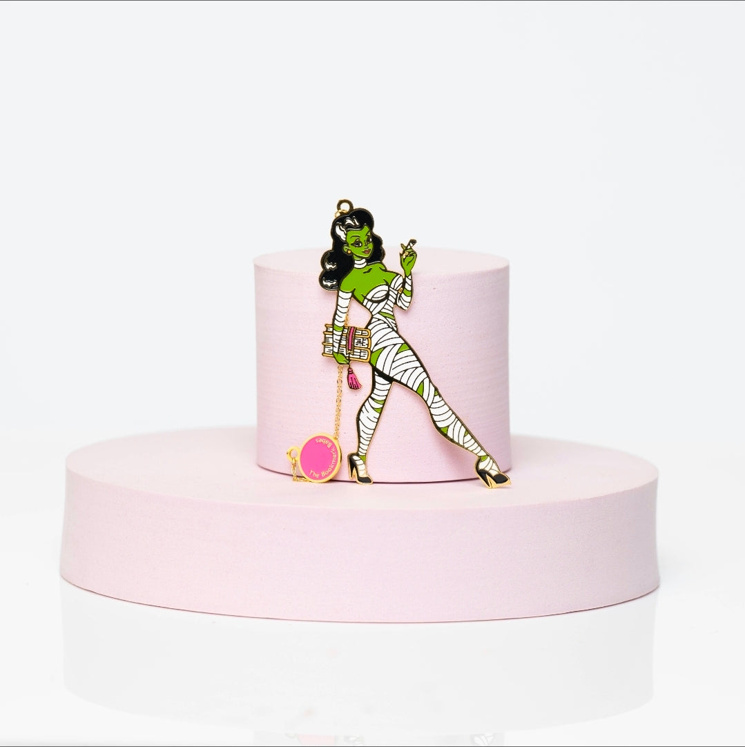 Green-skinned enamel bookmark character wrapped in mummy bandages, smoking a joint while holding a pink potion bottle. Wearing gold heels with a dangling pink charm and gold chain, posed on a soft pink pedestal.