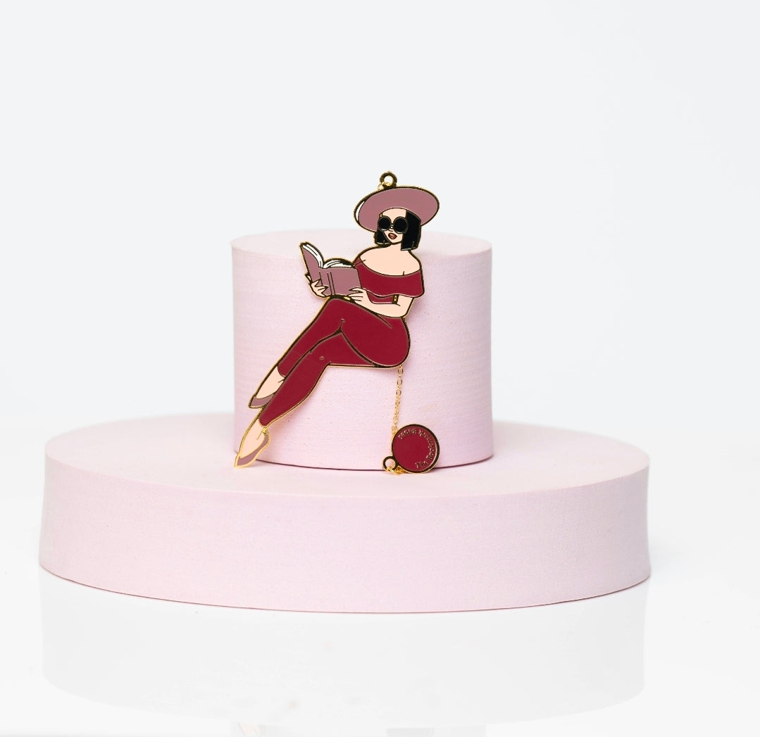 Enamel bookmark featuring Portia, a stylish Black woman in a red catsuit, reading a book with sunglasses and a wide-brim hat. Includes gold chain and charm, displayed on a pink pedestal.