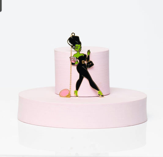 Enamel bookmark of Stack, a green-skinned woman in a sleek black catsuit, holding a pink book and handbag. Features gold detailing and a pink charm, displayed on a pink pedestal.
